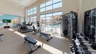 This fitness center is designed to support a comprehensive workout experience within a spacious, single-level layout. The main exercise area is defined by soaring ceilings and an impressive wall of floor-to-ceiling windows that frame views of an outdoor pool and surrounding amenity spaces, flooding the room with abundant natural light throughout the day. Neutral-toned carpet flooring and large wall mirrors visually expand the space while contributing to a clean, modern aesthetic.

The cardio and strength zone is thoroughly equipped with treadmills, stationary bikes, ellipticals, cable machines, and a variety of selectorized strength equipment, offering options for both machine-based and free-weight training. Adjustable benches and additional cable systems are positioned to maximize functionality across the floor.

A separate studio area provides space for group or individual exercise, featuring a bold geometric accent wall in teal and white, a wall-mounted fitness display screen, a rack of kettlebells, stackable aerobic steps, and a ball rack stocked with medicine and stability balls. Posted studio rules ensure an organized, shared environment. Together, these zones create a well-rounded fitness facility suited for a wide range of training needs.