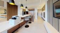 Located at 222 M Street in Washington, DC, this single-floor residential lobby showcases a sleek, contemporary design that seamlessly blends functionality with modern aesthetics. The space is anchored by dramatic floor-to-ceiling windows that flood the interior with abundant natural light while offering clear views of the surrounding streetscape, creating an open and inviting atmosphere.

The lounge area features a generously sized, upholstered sectional sofa accented with decorative throw pillows in muted earth and teal tones, complemented by sculptural side tables in contrasting finishes. Light-toned tile flooring extends throughout, enhancing the bright, airy feel of the space.

Moving deeper into the lobby, a well-appointed concierge station takes center stage, finished in warm walnut wood cabinetry paired with a clean white work surface. Workstations equipped with desktop computers and printing equipment are positioned beneath striking matte-black pendant lights with gold interiors, adding a refined industrial touch. Adjacent to the concierge area, a built-in mailroom wall provides organized resident mail access. Vertical wood slat wall paneling adds texture and warmth throughout, tying the design elements together cohesively. The overall ambiance is polished, professional, and welcoming—reflecting the quality and character of a premier Washington, DC address.