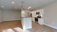 This well-appointed single-floor residence offers a thoughtfully designed open-concept layout that seamlessly connects the kitchen, dining, and living areas. The kitchen is a standout feature, showcasing crisp white shaker cabinetry, dark granite countertops, a central island with pendant lighting, and a full suite of stainless steel appliances including a refrigerator, electric range, built-in microwave, and dishwasher. Light hardwood-style flooring runs throughout the main living areas, reflecting natural light that pours in through large windows and a sliding glass door that opens to a private balcony. The living area is generously proportioned and benefits from excellent natural light, creating a bright and airy atmosphere. Two bedrooms are finished with neutral carpet, ceiling fans, and windows with natural wooded views. The unit includes two bathrooms: one featuring a tiled tub-shower combination with a white vanity, and a second with a curved glass-enclosed walk-in shower, a window for natural light, and tile flooring. Throughout the space, neutral gray-toned walls, recessed lighting, and clean white trim contribute to a cohesive, modern aesthetic that is both functional and inviting.