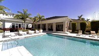 This single-story property showcases a sophisticated outdoor living environment centered around a pristine rectangular swimming pool with crystal-clear turquoise water. The pool area is framed by a refined covered pavilion supported by stately stacked-stone columns, offering a shaded retreat that seamlessly connects indoor and outdoor spaces. Steps lead up from the pool deck to the pavilion, enhancing the sense of elevated, resort-style living.

The exterior architecture blends contemporary and classical influences, featuring clean lines, a neutral color palette, and a pitched roof with dark tiles. Large openings within the pavilion provide unobstructed views and allow natural light and fresh air to flow freely throughout the space.

Surrounding the pool, the outdoor deck offers ample space for lounge seating, with stylish white outdoor furniture arranged for relaxation and entertaining. Mature palm trees and manicured tropical greenery frame the property, reinforcing the warm, sun-drenched atmosphere. The overall ambiance is one of refined leisure, combining elegant architectural detailing with lush landscaping for an inviting, upscale outdoor experience.