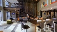 This dynamic two-story space makes an immediate impression with its soaring double-height ceilings and an architecturally distinctive open-riser staircase framed by a bold vertical slatted wood screen. The design seamlessly blends industrial and contemporary aesthetics, with clean lines, neutral tones, and carefully chosen accent pieces that create a refined yet welcoming atmosphere.

The main floor centers around a generous open-plan lounge and reception area. A communal bar-height table with sleek modern seating anchors one side of the space, offering a functional yet stylish gathering point. The lounge area is furnished with a plush sectional sofa in charcoal grey, accented with colorful throw pillows, alongside woven accent chairs that add warmth and texture. Contemporary coffee tables complete the seating arrangement, encouraging conversation and relaxation.

Polished large-format flooring runs throughout, reflecting the ambient recessed lighting that brightens the interior uniformly. Colorful artwork adorns the upper walls, adding vibrancy against the muted grey palette. The second floor, accessible via the statement staircase, features an open mezzanine with metal railings that overlooks the main floor below, reinforcing the sense of openness and vertical scale. The overall ambiance is modern, polished, and thoughtfully curated.