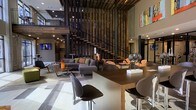 This dynamic two-story space makes an immediate impression with its soaring double-height ceilings and an architecturally distinctive open-riser staircase framed by a bold vertical slatted wood screen. The design seamlessly blends industrial and contemporary aesthetics, with clean lines, neutral tones, and carefully chosen accent pieces that create a refined yet welcoming atmosphere.

The main floor centers around a generous open-plan lounge and reception area. A communal bar-height table with sleek modern seating anchors one side of the space, offering a functional yet stylish gathering point. The lounge area is furnished with a plush sectional sofa in charcoal grey, accented with colorful throw pillows, alongside woven accent chairs that add warmth and texture. Contemporary coffee tables complete the seating arrangement, encouraging conversation and relaxation.

Polished large-format flooring runs throughout, reflecting the ambient recessed lighting that brightens the interior uniformly. Colorful artwork adorns the upper walls, adding vibrancy against the muted grey palette. The second floor, accessible via the statement staircase, features an open mezzanine with metal railings that overlooks the main floor below, reinforcing the sense of openness and vertical scale. The overall ambiance is modern, polished, and thoughtfully curated.