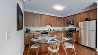 This single-floor space is designed with both functionality and comfort in mind. The kitchen is a standout feature, outfitted with rich wood cabinetry that extends to the ceiling, offering generous storage throughout. Granite countertops run along the full length of the kitchen, providing ample workspace, while stainless steel appliances — including a dishwasher, electric range, and microwave — deliver a modern, polished look. Crown molding adds a refined architectural touch along the ceiling line, enhancing the overall elegance of the space. Warm hardwood flooring flows seamlessly through the kitchen and into the adjacent dining area, creating a cohesive and inviting atmosphere. The dining area is centered around a round glass-top table surrounded by contemporary wood chairs, making it an ideal setting for both casual meals and entertaining guests. Overhead, a flush-mount ceiling light provides bright, even illumination throughout. The layout is thoughtfully arranged to maximize flow between the kitchen and dining areas, making everyday living both practical and comfortable. Overall, the space strikes a balance between classic design elements and modern finishes.