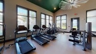 This fitness center is a thoughtfully designed, single-floor workout facility that combines functionality with motivating aesthetics. The space is immediately distinguished by its soaring vaulted ceilings, accented by a bold forest green upper trim that adds a striking visual contrast to the neutral-toned walls. A large ceiling fan anchors the room overhead, ensuring comfortable airflow throughout workouts.

The cardio zone is well-appointed with multiple treadmills, elliptical trainers, and a stationary bike, all positioned along a wall of generously sized windows. These large, dark-framed windows flood the space with natural light and offer views of the surrounding outdoor environment, creating an open, energizing atmosphere.

On the opposite end, the strength training area is equally impressive. A full Smith machine with weight plates and an adjustable bench serves as the centerpiece, complemented by a rack of dumbbells and additional cable resistance machines. The durable, light-toned flooring throughout is well-suited for high-traffic fitness use.

A standout feature of the space is the large-scale motivational mural that spans one full wall, depicting fitness-themed illustrations alongside an inspirational quote, reinforcing the facility's energetic and performance-driven character. Together, these elements create a comprehensive, welcoming fitness environment designed to support a full range of workout routines.