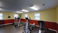 Located at 100 Tanglewood Dr in Southern Pines, NC, this fitness center is a single-floor space designed to support cardiovascular exercise and general wellness. The room features bold two-tone walls with a deep red lower half and warm yellow upper half, creating an energetic atmosphere suited for physical activity. Large flush-mounted fluorescent light panels line the dark ceiling, ensuring bright, even illumination throughout the space. Multiple windows with horizontal blinds allow natural light to filter in, contributing to an open and airy feel. The flooring consists of dark rubber matting, a practical and durable surface designed to withstand heavy equipment and high-traffic use. The fitness center is equipped with several treadmills, elliptical trainers, and stationary bikes, all positioned to allow comfortable use and adequate spacing between machines. A small piece of wall art adds a subtle decorative touch near one of the windows. The layout is straightforward and functional, with the equipment arranged to maximize available floor space while maintaining ease of movement throughout the room.