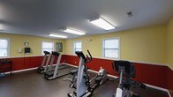 Located at 100 Tanglewood Dr in Southern Pines, NC, this fitness center is a single-floor space designed to support cardiovascular exercise and general wellness. The room features bold two-tone walls with a deep red lower half and warm yellow upper half, creating an energetic atmosphere suited for physical activity. Large flush-mounted fluorescent light panels line the dark ceiling, ensuring bright, even illumination throughout the space. Multiple windows with horizontal blinds allow natural light to filter in, contributing to an open and airy feel. The flooring consists of dark rubber matting, a practical and durable surface designed to withstand heavy equipment and high-traffic use. The fitness center is equipped with several treadmills, elliptical trainers, and stationary bikes, all positioned to allow comfortable use and adequate spacing between machines. A small piece of wall art adds a subtle decorative touch near one of the windows. The layout is straightforward and functional, with the equipment arranged to maximize available floor space while maintaining ease of movement throughout the room.