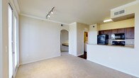 This single-floor residence features a 1-bedroom, 1-bathroom layout designed for efficient use of space. The open living area is finished with neutral-toned carpeting and crisp white walls accented by crown molding, creating a clean and inviting atmosphere. Track lighting along the ceiling provides versatile illumination throughout the main living space.

The kitchen is positioned adjacent to the living area and is partially separated by a raised breakfast bar counter, offering a natural division while maintaining an open feel. Dark cabinetry contrasts with light countertops, and a black refrigerator anchors the kitchen's appliance setup. The hard flooring in the kitchen adds a practical contrast to the carpeted living space.

An arched doorway leads from the living area toward the bedroom, lending an architectural detail that adds character to the otherwise straightforward layout. A glimpse through the corridor reveals what appears to be an in-unit washer/dryer area, adding notable convenience. The neutral color palette throughout the unit contributes to a calm, cohesive ambiance well-suited for a range of furnishing styles.