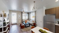 This single-story residence offers a thoughtfully designed layout with a cohesive modern aesthetic. The open-concept living and dining area serves as the heart of the home, featuring wood-look plank flooring, neutral-toned walls, and multiple windows dressed with blinds and grommet-panel curtains that invite an abundance of natural light. A ceiling fan overhead and a decorative chandelier above the dining area add functional yet stylish lighting elements. A sliding glass door provides direct access to the exterior, further enhancing the bright, airy feel of the space.

The kitchen is efficiently designed with warm wood-toned cabinetry extending to the ceiling for ample storage. Stainless steel appliances, including a refrigerator, range, over-the-range microwave, and dishwasher, are paired with light countertops and a single-basin sink positioned beneath a window. A white dining table with cognac-toned chairs connects the kitchen to the living area seamlessly.

The unit includes two bedrooms. The primary bedroom features a deep charcoal accent wall, warm ambient sconce lighting, a walk-in closet, and wood-look flooring. The secondary bedroom offers a ceiling fan, a large window, and a more neutral palette. Both rooms maintain a calm, restful atmosphere consistent with the overall design of the home.