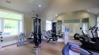 This fitness room offers a well-appointed workout environment designed to support a comprehensive exercise routine. The space is characterized by soaring vaulted ceilings that create an open, airy atmosphere, while recessed lighting and natural light from multiple windows keep the room bright and inviting. The neutral-toned walls and carpeted flooring contribute to a clean, comfortable setting.

The room is equipped with a multi-station cable weight machine, a weight bench, and various strength-training equipment, providing options for a full-body workout. A Life Fitness cardio machine is positioned toward the front of the space, adding versatility for aerobic training. A water cooler is conveniently placed along one wall, ensuring hydration is readily accessible during workouts.

A large wall mirror spans a significant portion of the room, visually expanding the space and allowing users to monitor their form during exercise. The overall layout is thoughtfully organized, ensuring ease of movement between equipment. This fitness room combines functionality with a welcoming ambiance, making it a practical and enjoyable space for daily exercise.
