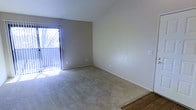 This single-floor space features a open living area defined by neutral beige walls and soft wall-to-wall carpeting that extends throughout most of the room, transitioning to hardwood flooring near the entryway. The ceiling rises to a vaulted peak, adding a sense of vertical spaciousness that enhances the room's overall openness. A sliding glass door fitted with vertical blinds serves as the primary source of natural light, allowing sunlight to filter into the space and illuminate the interior during daytime hours. The door likely leads to an outdoor area such as a patio or balcony, offering potential views of the surrounding exterior. The walls are smooth and neutral in tone, providing a clean, versatile backdrop suitable for a variety of interior styles. Electrical outlets and switch plates are positioned at standard heights along the walls. The combination of vaulted ceilings, natural light from the sliding door, and the open floor plan contributes to an airy, uncluttered ambiance throughout the space.
