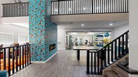 This well-appointed two-floor amenity space offers a generous and thoughtfully designed environment across two levels connected by a sleek staircase with bold black metal railings. The ground floor immediately captivates with its dramatic floor-to-ceiling mosaic tile fireplace — a vivid composition of teal, red, black, and white fragments that serves as the architectural centerpiece of the space. Light wood-style flooring runs throughout, complementing the clean, neutral walls and creating a warm, modern aesthetic. A large conference or activity table anchors this main gathering area, which also features seating nooks outfitted with teal accent chairs, adding pops of color. The exercise room is enclosed by full glass walls, offering a transparent view of the well-equipped fitness area. Inside, cardio machines including treadmills, ellipticals, and a stationary bike are arranged alongside strength equipment and a full-wall mirror that amplifies the space. A ceiling fan and checkered tile flooring complete the functional workout environment. Ascending to the upper level, soaring vaulted ceilings with exposed black beams create an airy, open atmosphere. A comfortable lounge area with teal and orange accent chairs invites relaxed gathering, while large windows fill the space with natural light. A sectional sofa area offers additional casual seating, rounding out this versatile upper floor.