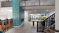 This well-appointed two-floor amenity space offers a generous and thoughtfully designed environment across two levels connected by a sleek staircase with bold black metal railings. The ground floor immediately captivates with its dramatic floor-to-ceiling mosaic tile fireplace — a vivid composition of teal, red, black, and white fragments that serves as the architectural centerpiece of the space. Light wood-style flooring runs throughout, complementing the clean, neutral walls and creating a warm, modern aesthetic. A large conference or activity table anchors this main gathering area, which also features seating nooks outfitted with teal accent chairs, adding pops of color. The exercise room is enclosed by full glass walls, offering a transparent view of the well-equipped fitness area. Inside, cardio machines including treadmills, ellipticals, and a stationary bike are arranged alongside strength equipment and a full-wall mirror that amplifies the space. A ceiling fan and checkered tile flooring complete the functional workout environment. Ascending to the upper level, soaring vaulted ceilings with exposed black beams create an airy, open atmosphere. A comfortable lounge area with teal and orange accent chairs invites relaxed gathering, while large windows fill the space with natural light. A sectional sofa area offers additional casual seating, rounding out this versatile upper floor.