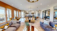 This contemporary two-story community lounge space spans a generous footprint across its upper level, offering a well-appointed and thoughtfully designed gathering environment. Entry is made through a dramatic double-door foyer featuring soaring ceilings, light wood flooring, geometric pendant lighting, and a modern metal staircase railing — setting an immediately sophisticated tone.

The main lounge level opens into a bright, expansive great room bathed in natural light from floor-to-ceiling windows and glass doors. A warm terracotta accent wall anchors one end of the space, complemented by an eclectic mix of plush seating in earthy gold and blue tones arranged around area rugs. A sleek wall-mounted fireplace topped with a large-screen television serves as a central focal point, flanked by curated artwork and decorative cabinetry.

Toward the back, a casual dining zone features round tables with cognac leather chairs beneath elegant chandelier lighting, adjacent to a kitchenette with dark wood cabinetry and a marble-style backsplash. Glass-partitioned rooms with black-framed doors provide flexible private spaces off the main lounge.

The highlight of the space is a covered outdoor terrace accessible via multiple glass doors, offering expansive panoramic views of the surrounding landscape. Furnished with outdoor seating and ceiling fans, this terrace provides a comfortable extension of the interior living area for year-round enjoyment.