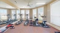 Located at 5400 Sycamore School Rd in Fort Worth, Texas, this dedicated fitness room offers a practical and well-organized space for residents to pursue their health and wellness goals. The single-floor layout is thoughtfully designed to maximize functionality, with a carpeted floor that provides a comfortable surface for a variety of workout activities.

The room is outfitted with an impressive selection of equipment, including multiple elliptical machines, a stationary recumbent bike, an upright cycle, and a rowing-style ab bench. A freestanding dumbbell rack with an assortment of weights, resistance tools, and a balance dome round out the free weights area, providing options for strength and stability training.

Floor-to-ceiling mirrors line one wall, visually expanding the space and allowing users to monitor their form during exercise. Multiple large windows fitted with horizontal blinds flood the room with natural light while offering views of the surrounding outdoor area. Ceiling fans provide continuous air circulation, enhancing comfort during workouts, while overhead fluorescent lighting ensures the space remains bright at all hours.

The warm, neutral-toned walls and clean white trim contribute to an inviting and motivating atmosphere. A direct exterior door provides convenient access to the outdoors, and posted workout guides on the wall serve as a helpful resource for fitness routines.