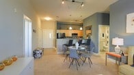 This single-floor residence offers a well-designed open-concept layout that seamlessly connects the living, dining, and kitchen areas into one cohesive space. The kitchen is outfitted with warm maple cabinetry, dark appliances, and a raised breakfast bar with counter seating, illuminated by elegant pendant lights that hang above. Track lighting along the ceiling provides additional ambient illumination throughout the main living area. The dining area sits at the heart of the space, centered around a glass-top round table surrounded by modern gray chairs, creating an inviting setting for everyday meals or entertaining. The flooring transitions from tile near the entryway to plush carpet in the living areas, adding warmth and texture underfoot. Accent walls in a muted teal-gray tone add depth and a contemporary aesthetic to the interior. A hallway near the front entry leads to additional rooms, while a glimpse of a bathroom is visible in the background, suggesting a thoughtful and functional floor plan. The overall ambiance is modern and comfortable, with a neutral color palette, tasteful furnishings, and carefully considered lighting that together create a welcoming and livable environment.