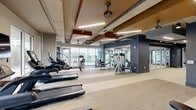 Located at 500 W Trade St in the heart of Charlotte, NC, this generously sized fitness center offers a comprehensive, well-appointed exercise facility spread across a single floor. The space is thoughtfully designed with a modern industrial aesthetic, featuring exposed ductwork, structural beams with warm wood-toned accents, and a coffered ceiling with integrated linear lighting that creates a bright, energizing atmosphere.

The facility is organized into distinct functional zones. A dedicated cardio area lines the perimeter walls, equipped with treadmills, ellipticals, stair climbers, and stationary bikes, all positioned in front of expansive floor-to-ceiling windows that frame views of the surrounding Charlotte cityscape and greenery. A strength training zone features a robust selection of selectorized machines, including Precor equipment, arranged across open, light-colored flooring. A free weights section provides a full rack of dumbbells accompanied by adjustable benches and rubber mat flooring for safety and comfort.

A glass-enclosed studio or flex space offers additional room for stretching, classes, or focused training, featuring mirrored walls and its own television display. Wall-mounted flat-screen TVs are positioned throughout the facility. The entrance corridor, finished in deep navy tones with large glass panel walls, provides a polished transition into the workout areas. Ceiling fans and climate control systems ensure comfort throughout.