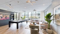 This thoughtfully designed amenity lounge offers a well-rounded social and recreational experience within a single, open-plan space. Large floor-to-ceiling windows and glass walls span the perimeter, flooding the interior with natural light and framing views of the surrounding urban landscape and outdoor terrace. Light hardwood flooring runs throughout, lending warmth and continuity to the space.

At the center of the room, a sleek pool table with a white felt surface serves as a focal point for recreation. Nearby, a built-in dark wood entertainment wall with backlit shelving and bold artwork adds a sophisticated, lounge-like atmosphere. A high-top dining table with leather chairs provides a casual setting for games or conversation.

The lounge area features a generously sized blush sectional sofa and plush accent chairs arranged around a round coffee table, creating an inviting space for relaxation. Decorative botanical wallpaper in soft blue-grey tones lines accent walls, adding texture and visual interest. A vintage-style arcade game cabinet tucks neatly into a corner, contributing a playful element. Designer sputnik-style pendant lighting and recessed fixtures complete the modern aesthetic, making this space equally suited for lively gatherings and quiet leisure.