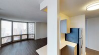 This single-floor unit is designed with a clean, contemporary aesthetic that maximizes both function and style. The open living area is anchored by expansive floor-to-ceiling windows that frame sweeping urban skyline views, flooding the space with natural light and creating a strong visual connection to the city below. Dark hardwood flooring runs throughout, providing a grounded contrast to the bright, neutral-toned walls and white textured ceiling.

A structural column subtly defines the transition between the living area and a dedicated built-in workspace. The work nook features a sleek, wall-mounted desk with an L-shaped white countertop, complemented by modern grey cabinetry above and a dark accent backsplash panel that adds visual depth. Additional lower cabinetry provides practical storage, making this area both organized and efficient.

The overall layout is thoughtfully arranged to balance open living space with purposeful functional zones. The minimalist design language, neutral color palette, and high-rise positioning combine to create an environment that feels bright, airy, and distinctly urban in character.