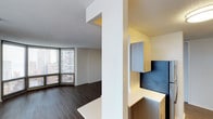 This single-floor unit is designed with a clean, contemporary aesthetic that maximizes both function and style. The open living area is anchored by expansive floor-to-ceiling windows that frame sweeping urban skyline views, flooding the space with natural light and creating a strong visual connection to the city below. Dark hardwood flooring runs throughout, providing a grounded contrast to the bright, neutral-toned walls and white textured ceiling.

A structural column subtly defines the transition between the living area and a dedicated built-in workspace. The work nook features a sleek, wall-mounted desk with an L-shaped white countertop, complemented by modern grey cabinetry above and a dark accent backsplash panel that adds visual depth. Additional lower cabinetry provides practical storage, making this area both organized and efficient.

The overall layout is thoughtfully arranged to balance open living space with purposeful functional zones. The minimalist design language, neutral color palette, and high-rise positioning combine to create an environment that feels bright, airy, and distinctly urban in character.