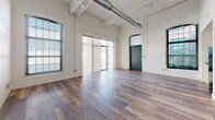 Located at 166 Valley St in Providence, Rhode Island, this single-floor loft-style unit showcases the character of an industrial conversion with thoughtfully updated modern amenities. The expansive main living area immediately impresses with soaring ceilings, exposed spiral ductwork, and painted wood-plank ceiling details that speak to the building's historic origins. Rich, wide-plank hardwood flooring flows throughout the open space, complemented by whitewashed brick walls that add warmth and texture. Generous arched and multi-pane windows flood the interior with abundant natural light, enhancing the airy, open atmosphere. The kitchen is well-appointed with granite countertops, a stainless steel sink, gas range, dishwasher, and light wood cabinetry, with an exposed brick accent wall adding industrial charm. A dedicated laundry area with stacked washer and dryer units adds everyday convenience. The bathroom features a full tub and shower combination with clean white tile surround, granite-topped vanity, and light tile flooring. A bold green arched doorway adds a distinctive architectural accent, nodding to the building's storied past. The overall ambiance blends historic industrial character seamlessly with contemporary living comforts.