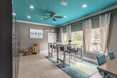 This single-floor communal space makes an immediate design statement with its striking teal-painted ceiling, which anchors the room's bold, contemporary aesthetic. Light-toned porcelain tile flooring spans the entire space, complementing the warm gray walls and crisp white trim throughout. A ceiling fan with recessed lighting keeps the room comfortable and well-lit.

The main gathering area features a high-top dining table paired with upholstered bar-height chairs, set atop a colorful chevron-patterned area rug in teal, yellow, and gray tones. Multiple large windows dressed with geometric-patterned curtains and gray roller shades allow generous natural light to filter in, offering outdoor views of surrounding greenery and what appears to be a pool area.

A dedicated work zone includes built-in desk surfaces along the wall, equipped with teal ergonomic task chairs, making the space functional for both collaborative and focused work. Bold abstract artwork adorns the walls, reinforcing the space's modern, curated feel.

Through French doors, the space opens into a broader lounge and common area featuring soaring ceilings, a seating arrangement with sofas and accent rugs, and a glimpse of a fully equipped kitchen beyond, suggesting this is part of a larger amenity or clubhouse facility.