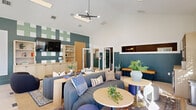 This thoughtfully designed living room offers a spacious, open-plan layout that seamlessly blends lounge and collaborative dining areas under soaring vaulted ceilings. The architectural design is defined by crisp white upper walls contrasted with bold teal wainscoting, creating a modern yet inviting atmosphere throughout the space.

The lounge area anchors the room with a generously sized charcoal channeled sofa, complemented by curved accent chairs, leather ottomans, and layered area rugs that add warmth and texture. A sculptural matte-black sputnik chandelier serves as a striking focal point overhead, while recessed skylights and flush-mount linear fixtures ensure the space is well-lit at all times.

A large natural wood communal table with matching black chairs defines the dining and collaborative zone, ideal for group gatherings or work sessions. Built-in wood shelving units provide both display and storage, and a dual-zone beverage refrigerator adds a convenient amenity for residents.

Natural light pours in through multiple windows dressed with woven bamboo shades, offering views of the surrounding outdoors. Light wood-toned flooring runs throughout, tying the various zones together cohesively. An accent wall featuring a graphic teal-and-white geometric pattern adds an artistic touch, reinforcing the space's contemporary, community-centered character.
