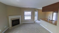 This single-floor space centers around a comfortable living area finished with wall-to-wall carpeting in a neutral tone, creating a warm and cohesive atmosphere throughout. A standout feature is the fireplace framed by a classic white mantel and tiled surround, positioned along one wall and serving as a natural focal point for the room.

Natural light fills the space through a large double-hung window that offers outdoor views of surrounding trees and greenery, while a sliding glass door nearby provides additional light and potential outdoor access. The walls are finished in a warm beige tone, complementing the light-colored carpet and white trim work.

Adjacent to the main living area, a transitional space leads to what appears to be a kitchen, distinguished by a partial view of dark wood cabinetry and a countertop with a light stone finish. The kitchen is separated from the living area by a step up, adding subtle architectural definition between the two spaces.

The ceiling features a textured finish, and overall the layout offers an open, functional flow between the living and kitchen areas, making efficient use of the available space.