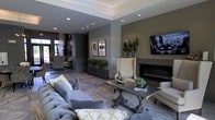 This single-floor space showcases a refined and contemporary interior design, centered around an expansive lounge area that exudes elegance and comfort. The room features a striking tray ceiling with recessed lighting, casting a warm, layered glow throughout the space. A sleek, wall-mounted linear fireplace serves as a focal point, framed by textured stone or tile surround and flanked by tall decorative plants that add a natural, organic element to the sophisticated aesthetic.

The lounge is furnished with plush, tufted gray sofas accented by yellow and green throw pillows, complemented by striped wingback chairs that add a classic touch to the modern setting. A wooden trestle coffee table anchors the seating arrangement, topped with curated greenery and books. Framed black-and-white artwork adorns the walls, contributing to the space's gallery-like character.

Wide-plank flooring in a warm wood tone extends throughout, and large windows or glass doors at the far end invite natural light deep into the interior, offering views of lush outdoor greenery. The overall ambiance is polished and inviting, balancing modern design sensibilities with warm, livable details that make the space both visually impressive and functionally comfortable.