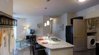This single-floor unit offers a thoughtfully designed open-concept layout that seamlessly connects the kitchen and living areas. The kitchen is a standout feature, showcasing a granite-topped peninsula that doubles as a breakfast bar with seating for multiple guests. A trio of pendant lights hangs above the counter, providing focused task lighting while adding a modern, decorative touch. The kitchen is equipped with dark appliances, including a full-size refrigerator, and features a built-in sink within the peninsula for added convenience. Dark wood-toned cabinetry provides ample storage. The flooring throughout is a warm hardwood, lending a cohesive and polished look to the space. The adjoining living area benefits from the open layout, creating an airy and connected feel. A wall-mounted shelving unit adds a decorative accent, while a floor lamp provides soft ambient lighting. Neutral wall tones throughout keep the space feeling bright and versatile, allowing the architectural details and fixtures to take center stage. The overall ambiance is contemporary and inviting, balancing functional design with clean, modern aesthetics suited for everyday living.