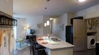 This single-floor unit offers a thoughtfully designed open-concept layout that seamlessly connects the kitchen and living areas. The kitchen is a standout feature, showcasing a granite-topped peninsula that doubles as a breakfast bar with seating for multiple guests. A trio of pendant lights hangs above the counter, providing focused task lighting while adding a modern, decorative touch. The kitchen is equipped with dark appliances, including a full-size refrigerator, and features a built-in sink within the peninsula for added convenience. Dark wood-toned cabinetry provides ample storage. The flooring throughout is a warm hardwood, lending a cohesive and polished look to the space. The adjoining living area benefits from the open layout, creating an airy and connected feel. A wall-mounted shelving unit adds a decorative accent, while a floor lamp provides soft ambient lighting. Neutral wall tones throughout keep the space feeling bright and versatile, allowing the architectural details and fixtures to take center stage. The overall ambiance is contemporary and inviting, balancing functional design with clean, modern aesthetics suited for everyday living.