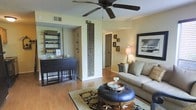 This single-floor residence offers a comfortable and cohesive living environment with a warm, inviting ambiance. The living area serves as the heart of the home, featuring hardwood-style flooring that extends throughout the space, lending a sense of continuity and warmth. A ceiling fan is centrally positioned overhead, providing both functional airflow and a classic design element. The living room is furnished with a neutral-toned sofa and a distinctive round leather ottoman that doubles as a coffee table, anchored by a patterned area rug that adds texture and visual interest. A decorative carved panel serves as an eye-catching architectural accent on the dividing wall, complementing the curated wall art and framed photography that adorn the surrounding walls. A table lamp adds soft ambient lighting near the seating area. Adjacent to the living space, a dining nook features a dark-toned drop-leaf table with matching stools, alongside a freestanding shelving unit ideal for display or storage. A hallway leads toward additional rooms, offering a sense of depth and privacy within the layout. The overall design blends casual comfort with decorative character, creating a lived-in yet stylish atmosphere throughout.