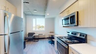 This single-floor unit showcases a thoughtfully designed, open-concept layout that seamlessly integrates the kitchen, dining, and living areas into one cohesive space. The interior is finished with light wood-look flooring throughout, contributing to a clean, contemporary aesthetic that is both functional and inviting.

The kitchen is outfitted with light wood cabinetry accented by gold hardware, stainless steel appliances including a refrigerator, electric range, microwave, and dishwasher, and ample counter space. An architectural highlight of the main living area is the partially exposed concrete ceiling, which lends an industrial-modern character to the space. A large window draws in generous natural light while framing urban views of the surrounding cityscape and greenery.

The living and dining areas provide room for a lounge seating arrangement and a compact bistro-style dining set, making efficient use of the available space. A dedicated in-unit stacked washer and dryer adds everyday convenience. The bathroom features a full tub-shower combination with clean white surrounds. Additional storage is provided via a shelved closet near the entryway, which also features a distinctive wood-paneled door.
