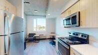 This single-floor unit showcases a thoughtfully designed, open-concept layout that seamlessly integrates the kitchen, dining, and living areas into one cohesive space. The interior is finished with light wood-look flooring throughout, contributing to a clean, contemporary aesthetic that is both functional and inviting.

The kitchen is outfitted with light wood cabinetry accented by gold hardware, stainless steel appliances including a refrigerator, electric range, microwave, and dishwasher, and ample counter space. An architectural highlight of the main living area is the partially exposed concrete ceiling, which lends an industrial-modern character to the space. A large window draws in generous natural light while framing urban views of the surrounding cityscape and greenery.

The living and dining areas provide room for a lounge seating arrangement and a compact bistro-style dining set, making efficient use of the available space. A dedicated in-unit stacked washer and dryer adds everyday convenience. The bathroom features a full tub-shower combination with clean white surrounds. Additional storage is provided via a shelved closet near the entryway, which also features a distinctive wood-paneled door.