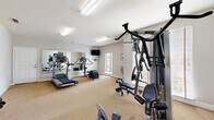This fitness center offers a practical and well-organized space designed to support a full range of workout routines. The room features neutral-toned walls, carpet flooring, and flush fluorescent lighting that ensures bright, even illumination throughout. Multiple windows fitted with blinds allow natural light to fill the space while maintaining privacy, contributing to an open and energizing atmosphere.

The facility is equipped with a diverse selection of cardio machines, including treadmills, an elliptical trainer, a stationary upright bike, and a rowing-style machine. For strength training, a multi-station cable machine — a Hoist brand unit — provides a variety of resistance exercises, complemented by a weight bench for additional training options. A wall-mounted flat-screen television is positioned for convenient viewing during workouts.

A large wall mirror along one side of the room visually expands the space and allows users to monitor their form. The room also includes a wall-mounted air conditioning and heating unit for year-round climate control. An emergency exit door with exterior access provides added safety and convenience. Informational signage and equipment guides are posted on the walls, making the space user-friendly. Overall, the fitness center delivers a clean, functional, and comfortable environment suited for daily exercise.