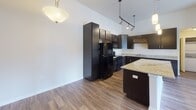 Located at 3110 Market Lane in Kenosha, Wisconsin, this single-floor residence offers a well-appointed layout designed for comfortable everyday living. The kitchen serves as the centerpiece of the home, showcasing rich dark cabinetry, light granite countertops, and a large center island that provides additional prep space and casual seating. Black appliances, including a refrigerator, range, and microwave, complement the bold cabinetry, while pendant lighting and a track lighting system illuminate the space with style. A decorative wine rack detail adds a refined touch above the upper cabinets. Wood-look flooring flows throughout the kitchen and main living areas, lending warmth and cohesion. In-unit stacked laundry is conveniently accessible just off the kitchen. The bedrooms feature soft carpeting and vaulted ceilings that lend an open, airy feel, with windows that allow natural light to filter in. Ceiling fans provide additional comfort. The bathrooms are finished with stone-look tile flooring and dark vanity cabinetry; one bathroom includes a soaking tub and shower combination, while the second offers a walk-in shower. Neutral wall tones throughout create a clean, versatile backdrop suited to a variety of décor styles.