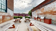 Located at 710 Greenville St in LaGrange, Georgia, this open-air courtyard offers a compelling blend of historic industrial character and functional outdoor gathering space. Enclosed on multiple sides by aged brick walls — some bearing layers of weathered paint that tell the story of the building's long history — the space exudes a raw, authentic ambiance that is both inviting and visually striking.

The courtyard features a spacious concrete patio accommodating multiple wooden picnic tables, making it well-suited for communal dining and group gatherings. String lights are strung overhead, providing a warm, festive atmosphere during evening hours. A dedicated outdoor kitchen area with built-in grills and cabinetry in a bold red finish offers practical cooking and entertaining capabilities.

Large multi-pane windows with green frames line the upper portion of the primary brick wall, allowing views into the adjoining building while adding architectural interest. A metal staircase is visible along one wall, providing access to an upper level. Mature landscaping with neatly trimmed shrubs and greenery softens the perimeter, and open sightlines to the surrounding neighborhood connect the space to its broader urban context. Overall, this courtyard is a distinctive, character-rich outdoor venue with strong industrial-historic appeal.