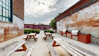 Located at 710 Greenville St in LaGrange, Georgia, this open-air courtyard offers a compelling blend of historic industrial character and functional outdoor gathering space. Enclosed on multiple sides by aged brick walls — some bearing layers of weathered paint that tell the story of the building's long history — the space exudes a raw, authentic ambiance that is both inviting and visually striking.

The courtyard features a spacious concrete patio accommodating multiple wooden picnic tables, making it well-suited for communal dining and group gatherings. String lights are strung overhead, providing a warm, festive atmosphere during evening hours. A dedicated outdoor kitchen area with built-in grills and cabinetry in a bold red finish offers practical cooking and entertaining capabilities.

Large multi-pane windows with green frames line the upper portion of the primary brick wall, allowing views into the adjoining building while adding architectural interest. A metal staircase is visible along one wall, providing access to an upper level. Mature landscaping with neatly trimmed shrubs and greenery softens the perimeter, and open sightlines to the surrounding neighborhood connect the space to its broader urban context. Overall, this courtyard is a distinctive, character-rich outdoor venue with strong industrial-historic appeal.