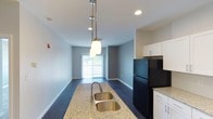 This well-appointed single-floor residence offers a thoughtfully designed open-concept layout that seamlessly connects the kitchen and living areas. The kitchen serves as a central hub of the space, anchored by a granite-topped island with a double stainless steel sink and elegant pendant lighting hanging above. White shaker-style cabinetry provides ample storage and pairs cleanly with the black refrigerator, creating a striking modern contrast. Recessed lighting supplements the natural illumination throughout. Rich, dark hardwood flooring runs throughout the main living area, adding warmth and a sense of cohesion to the overall design. The open living area extends naturally from the kitchen, bathed in natural light streaming through a large window or sliding glass door at the far end, offering a bright and airy atmosphere. The soft gray walls complement the flooring and cabinetry, lending a calm, neutral palette that suits a variety of interior styles. Clean lines, contemporary fixtures, and quality materials come together to create a functional and stylish living environment that feels both modern and welcoming.