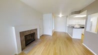 This single-floor residence offers a thoughtfully designed open-concept layout that seamlessly connects the main living area to the kitchen. The living room is anchored by a classic tile-surround fireplace, adding warmth and character to the space. Light hardwood flooring runs throughout, lending a clean, modern aesthetic that complements the bright, neutral-toned walls.

The kitchen is positioned adjacent to the living area and features white cabinetry, a peninsula counter with dark countertops, and a range hood overhead, making it both functional and visually distinct. The open sightlines between the kitchen and living room create a sense of spaciousness and ease of flow throughout the home.

Natural light filters into the space through well-placed windows, contributing to an airy and welcoming ambiance. The entryway, visible from the main living area, includes a white door that adds to the clean, minimalist aesthetic of the interior.

Overall, the space presents a practical and comfortable layout with notable design elements, including the tile fireplace surround and hardwood floors, that elevate the everyday living experience. The neutral palette throughout provides a versatile backdrop suitable for a variety of interior styles and furnishing choices.