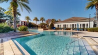 This outdoor amenity space is designed to evoke a resort-like atmosphere, centered around a generously sized swimming pool with exceptionally clear, blue-tinted water. The pool area features a multi-level layout, with raised stone-clad retaining walls adding architectural depth and visual interest to the deck. Blue lounge chairs are positioned throughout the sun-drenched pool surround, offering ample space for relaxation.

A separate spa is located adjacent to the main pool area, nestled within a more intimate setting framed by tall bamboo plantings, stone walls, and a wooden pergola structure that provides partial shade. Potted flowering plants and lush greenery enhance the tranquil ambiance of this zone.

The surrounding landscape is rich with mature palm trees, oak trees, and manicured garden beds filled with colorful flowering shrubs, contributing to a tropical aesthetic. Classic lamp posts punctuate the paved walkways, which are constructed from attractive interlocking pavers throughout the entire outdoor area.

A traditional-style single-story clubhouse with white-painted siding, large multi-pane windows, and a dark hip roof serves as the architectural anchor of the complex. The overall design of the space balances functionality with a welcoming, upscale outdoor environment.
