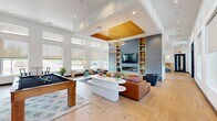 This thoughtfully designed single-floor community space offers a generous and versatile layout ideal for gathering, relaxing, and entertaining. The centerpiece of the space is a grand open-plan living area distinguished by soaring ceilings, a striking recessed wood-paneled tray ceiling, and cascading circular pendant lights finished in brushed gold. Light herringbone-patterned hardwood flooring flows throughout, anchoring the bright, airy atmosphere created by floor-to-ceiling windows fitted with roller shades. A dramatic accent wall clad in blue-gray tile houses a built-in fireplace, large flat-screen TV, and flanking wood shelving, providing a sophisticated focal point. Multiple seating arrangements with vibrant teal and cognac leather furnishings offer comfortable lounging, while a full-size pool table adds a recreational element. The adjacent kitchen and dining area features dual-tone cabinetry in navy and natural wood, a large marble-topped island with seating, textured tile backsplash, and sculptural pendant lighting beneath the wood tray ceiling. A secondary living room offers a more intimate setting, with warm wood wainscoting, patterned carpet, and a large-format display screen. A sun-drenched nook with panoramic windows and a bistro table completes this well-appointed space.