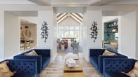 This single-floor community gathering space, known as the Town Center, offers a thoughtfully designed interior that blends contemporary style with warm, inviting details. The open-concept layout flows seamlessly between multiple lounge areas, defined by soaring vaulted ceilings with exposed wood beam accents and an abundance of natural light streaming through large, multi-pane windows.

The main lounge features shiplap-clad walls, a classic white fireplace with a wall-mounted television above, and flanking built-in shelving with decorative teal-toned accents. Eclectic furnishings — including patterned accent chairs, tufted navy velvet sofas, and a rich leather sectional — lend the space a curated, residential feel. A shuffleboard table adds a recreational focal point, making the area ideal for socializing and leisure.

Architectural columns frame the transition between seating zones, each adorned with striking black sculptural wall art. Wood-look tile flooring and bold patterned area rugs unify the space throughout.

The kitchenette area is equally impressive, featuring slate-blue cabinetry with gold hardware, a quartz countertop, and a vibrant mosaic-tile backsplash that serves as a bold artistic statement. Open wood shelving and a chalkboard welcome sign complete the charming, functional vignette. Together, these spaces create an engaging and versatile community environment.