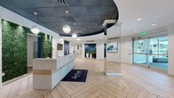Located at 5901 Montrose Rd in Rockville, Maryland, this single-floor leasing office lobby for Arrive is a thoughtfully designed commercial space that makes a strong first impression. The reception area serves as the centerpiece, featuring a sleek, modern desk set against a lush living green wall that adds a vibrant, natural contrast to the otherwise polished interior. A striking dark, curved ceiling medallion with globe pendant lights anchors the space overhead, drawing the eye upward and adding architectural depth.

The flooring throughout is a light-toned herringbone wood pattern that lends warmth and visual interest underfoot. A glass-enclosed private room adjacent to the main floor provides a more intimate setting for meetings or consultations. The lounge area is highlighted by a built-in navy upholstered bench positioned in front of large windows framed by teal-accented panels, offering views of the surrounding exterior and filling the space with natural light.

Curated artwork, potted greenery, and mirrored column accents contribute to the refined, welcoming ambiance. The automatic glass entry doors and open floor plan ensure ease of movement throughout the space. The overall design balances professionalism with approachability, making it a functional and visually compelling environment for welcoming prospective residents.
