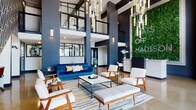The lobby of Flats Off Madison at 120 Malibu Dr in Clarksville, Tennessee, makes an immediate impression with its dramatic double-height ceilings and bold contemporary design. Towering dark structural columns frame the open volume, while cascading pendant chandeliers with elongated glass rods draw the eye upward and fill the space with warm, ambient light. A full-scale living green wall serves as the centerpiece backdrop, displaying the Flats Off Madison branding in luminous white lettering and infusing the space with a vibrant, natural element.

The main seating area is thoughtfully curated with mid-century modern furnishings, including a rich navy velvet sofa, warm wood-framed lounge chairs, and a sleek marble-top coffee table, all anchored by a colorful geometric area rug. Polished concrete floors run throughout, lending an urban, industrial character that complements the navy and dark gray color palette.

Near the entrance, an accent wall features an intricate gold geometric tile pattern, adding a sophisticated decorative detail alongside a console table and mirror. Large floor-to-ceiling windows and glass entry doors flood the space with natural light and offer clear sightlines to the exterior. A clean white reception desk provides a welcoming point of contact, and a dedicated coffee bar with a beverage cooler and sink offers a convenient amenity for residents and guests alike.