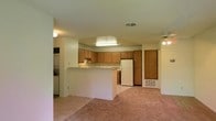 This single-story residence offers a practical and comfortable layout centered around an open-concept living and kitchen area. The kitchen is well-appointed with warm honey-toned wood cabinetry that extends to the ceiling, providing generous storage space. A built-in range hood and white refrigerator are seamlessly integrated into the design, while a peninsula breakfast bar creates a natural divide between the kitchen and the adjacent living space, ideal for casual dining or meal prep. Overhead, a skylight-style recessed panel light brightens the kitchen, complementing the ambient ceiling light fixtures throughout. The living area features soft wall-to-wall carpeting in a neutral tone, lending a cozy and inviting atmosphere to the space. A ceiling fan is also present, adding both function and comfort to the room. Along one wall, a stacked laundry unit is conveniently tucked into a dedicated alcove, maximizing the efficient use of space. The interior is finished in neutral tones throughout, creating a clean and versatile backdrop. HVAC vents are visible in the ceiling and walls, ensuring year-round climate comfort. Overall, this home delivers a cohesive, functional layout well-suited for everyday living.