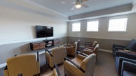 Located at 8101 Fonthill Court in Langhorne, Pennsylvania, this single-floor community space offers a comfortable and thoughtfully designed lounge environment. The media room serves as the focal point of the space, featuring a large wall-mounted flat-screen television positioned on a sleek, floating walnut media console with integrated storage. Multiple upholstered armchairs with dark wood frames are arranged in a semicircular configuration facing the screen, providing ample seating for group viewings or social gatherings. The room is finished with neutral gray walls accented by crisp white crown molding and a two-tone paint treatment that adds architectural interest. Carpeted flooring in a warm, muted tone contributes to a cozy atmosphere, while a ceiling fan with an integrated light fixture ensures comfortable air circulation throughout. Natural light filters in through multiple double-hung windows, brightening the space during daytime hours. The overall ambiance is clean, modern, and inviting, reflecting a well-maintained communal setting suited for relaxed entertainment and social interaction.