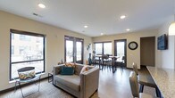 Located at 6634 Riverside Drive in Dublin, Ohio, this single-floor space offers an open, well-appointed interior designed for both comfort and functionality. The layout seamlessly connects a living area with a dining space, creating a cohesive environment well-suited for gatherings or everyday use.

The living area features a contemporary grey sofa accented with colorful throw pillows, positioned atop a patterned area rug over durable hardwood-style flooring. Recessed lighting in the white ceiling provides even illumination throughout, complementing the generous natural light that streams in through multiple large, dark-framed windows and glass-paneled doors.

The dining area accommodates a full table with seating for several guests and is positioned near additional glass doors that open directly to an outdoor deck or balcony, offering views of the surrounding landscape. Warm beige walls and dark trim details throughout lend the space a modern yet welcoming aesthetic. A decorative wall clock and subtle accent pieces add character without overwhelming the clean, functional design of the interior.
