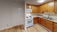 This single-floor unit offers a clean, modern aesthetic throughout, anchored by light-toned hardwood flooring that runs seamlessly across the living and bedroom areas. The open layout is complemented by crisp white walls and crown molding details that add a polished finish to the space.

The kitchen is a functional highlight, outfitted with warm honey-toned wood cabinetry extending floor to ceiling for ample storage. Light granite countertops provide generous prep space, while white appliances — including a gas range with over-the-range microwave and a full-size refrigerator — complete the well-equipped cooking area. A double-basin sink is positioned beneath efficient overhead lighting, making the kitchen both practical and inviting.

The bedroom is comfortably sized and features a ceiling fan with integrated lighting, a wall-mounted air conditioning unit for climate control, and a window dressed with vertical blinds that allow for adjustable natural light. The room's neutral palette and hardwood floors create a calm, airy atmosphere.

The bathroom is tastefully updated, showcasing large-format travertine-style tiles lining the walls of a deep soaking tub and shower combination. A frameless sliding glass door adds a contemporary touch, while a mosaic accent tile border provides refined detail. Overall, the space presents a tidy, move-in ready environment with consistent finishes throughout.