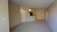 This single-floor space features an open-concept layout that connects the living area to the kitchen, creating a sense of spaciousness and easy flow between functional zones. The living area is finished with wall-to-wall carpeting in a neutral tone, contributing to a comfortable and understated ambiance. Recessed lighting is installed throughout, providing consistent, even illumination across the space.

The kitchen is partially separated from the main living area by a raised bar-height counter, which serves as both a visual divider and a practical surface for casual dining or food preparation. Upper cabinetry in a warm wood tone is visible beyond the counter, along with a dark-finish appliance that anchors the kitchen area. A wine rack detail adds a subtle design element above the cabinetry.

The entryway, accessible through a white interior door, transitions from tile flooring to the carpeted main living space, clearly delineating the entry zone. Walls throughout are painted in a soft, neutral tone, complementing the textured popcorn ceiling. A HVAC vent is visible, indicating a central air system. The overall design is functional and clean, with a straightforward layout suited for everyday living.