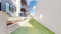 This outdoor space serves as a dedicated pet relief and recreation area situated along the ground level of a modern multi-story residential building. The elongated corridor-style layout is surfaced with well-maintained artificial turf, providing a clean and functional area for pets year-round. On one side, raised concrete retaining walls border tiered planting beds filled with young shrubs and ornamental plantings, adding a natural, landscaped aesthetic to the space. The opposite side features a smooth masonry wall that reflects natural sunlight, keeping the area bright and open to the sky above. At one end, concrete steps with metal handrails provide access from an upper walkway level, while a wooden privacy fence defines a secondary section of the turf area. The surrounding building facade combines brick, dark navy cladding, and glass-railed balconies rising several stories overhead, framing the space with a contemporary architectural character. Utility access points are flush-set within the turf, maintaining a clean visual appearance. The area benefits from open sky exposure, allowing ample natural light throughout the day, while the enclosed nature of the corridor provides a sense of privacy and security for pets and their owners.
